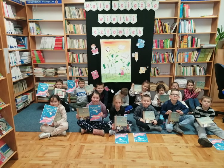 Na zdjęciu przedstawiona jest klasa pierwsza, która znajduje się w bibliotece szkolnej, siedem dziewczynek oraz siedmiu chłopców.  Wszyscy trzymają w rękach dyplomy oraz wyprawki wielkiego czytelnika „ Mała książka-wielki człowiek”.  Za nimi znajduję się dekoracja z dużym napisem „ Mała książka –wielki człowiek”, ilustracja przedstawiająca dziewczynkę podlewająca kwiaty oraz półki z książkami.